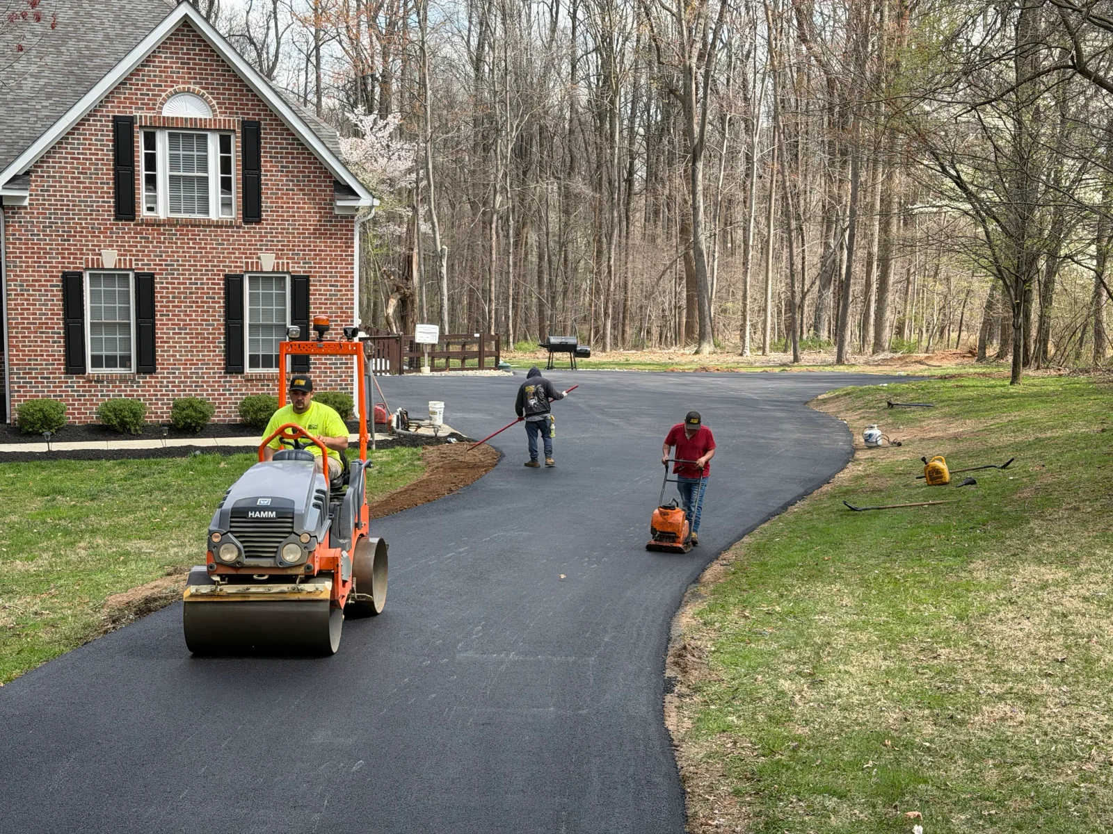 Crew compacting fresh asphalt driveway with HAMM roller, plate compactor, and rake beside brick home