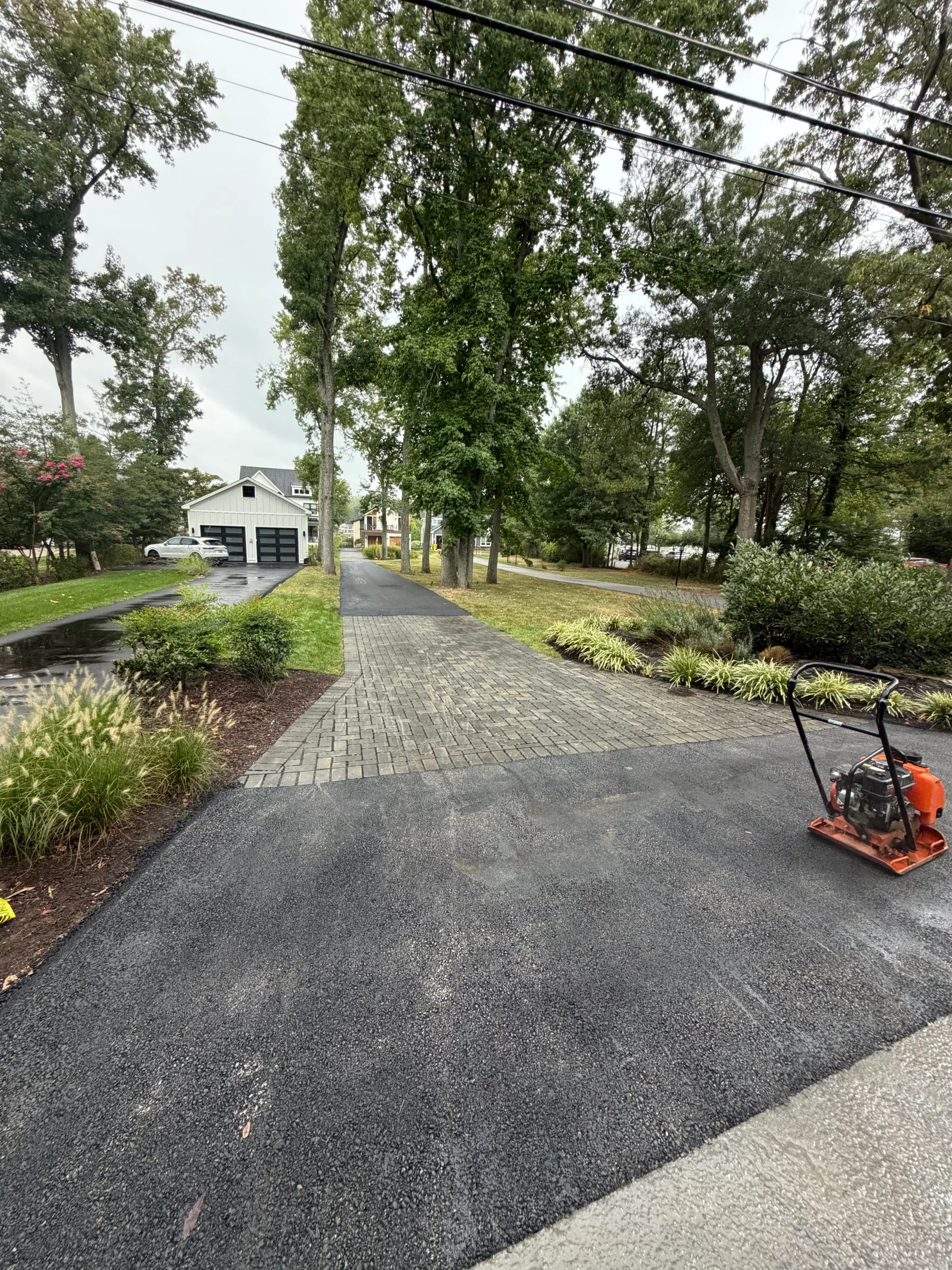 New black asphalt driveway with gray paver border, ornamental grass beds, and plate compactor on fresh asphalt