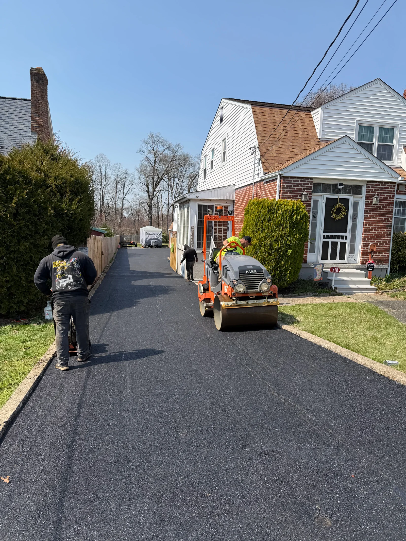 Freshly laid asphalt driveway being compacted by a HAMM tandem roller beside a brick home with curbed edges