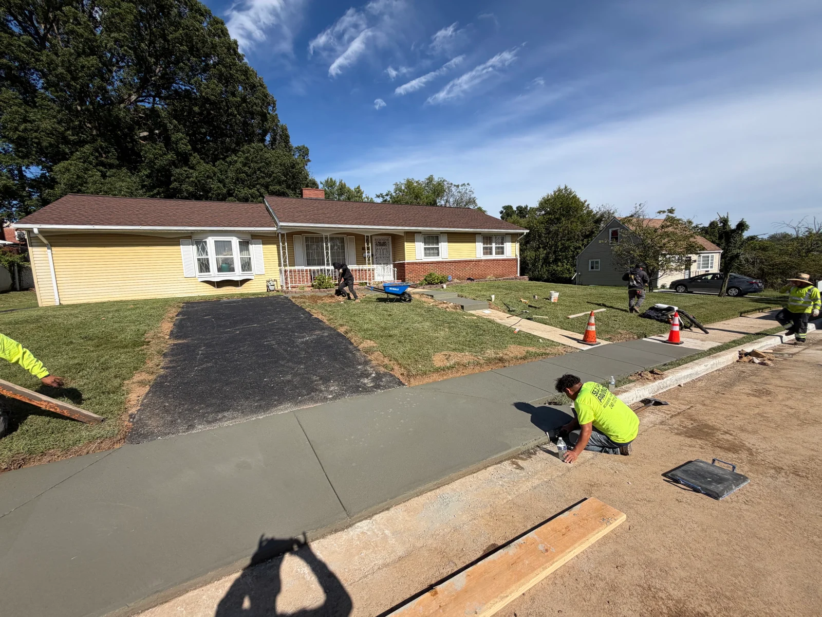 Freshly poured concrete sidewalk with wood forms beside a new asphalt driveway and sod installation at a residential front yard