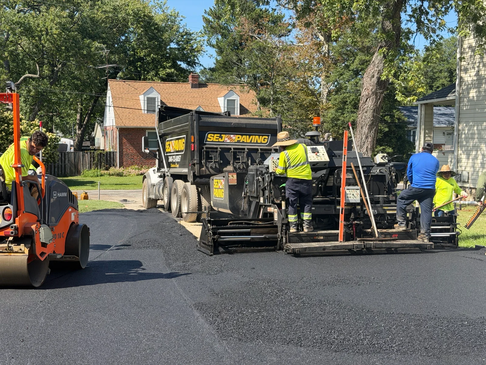 Crew paving fresh black asphalt with a tracked paver, dump truck, and Hamm roller on a residential street