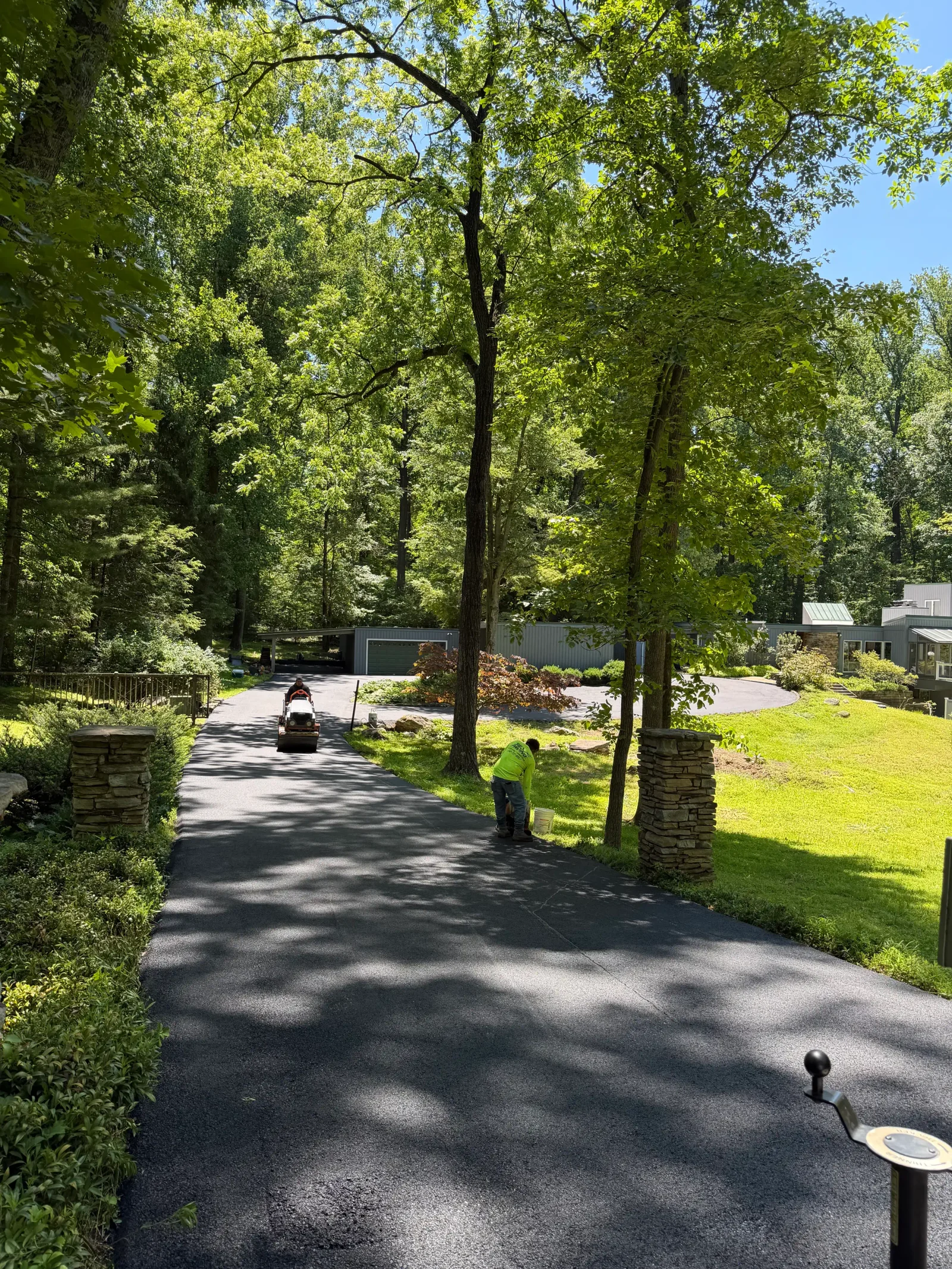 Freshly paved asphalt driveway with roller compactor, stacked stone entry pillars, and landscaped shrubs
