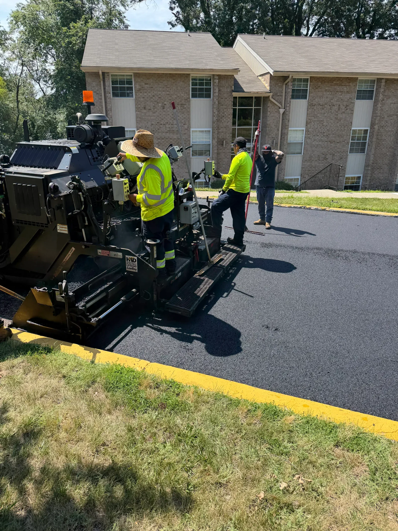 Crew operating tracked asphalt paver laying fresh asphalt along yellow-painted curb edge