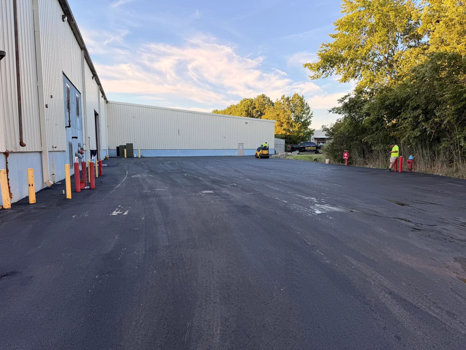 Newly paved black asphalt lot beside a metal industrial building with yellow and red bollards and a yellow roller compacting the surface