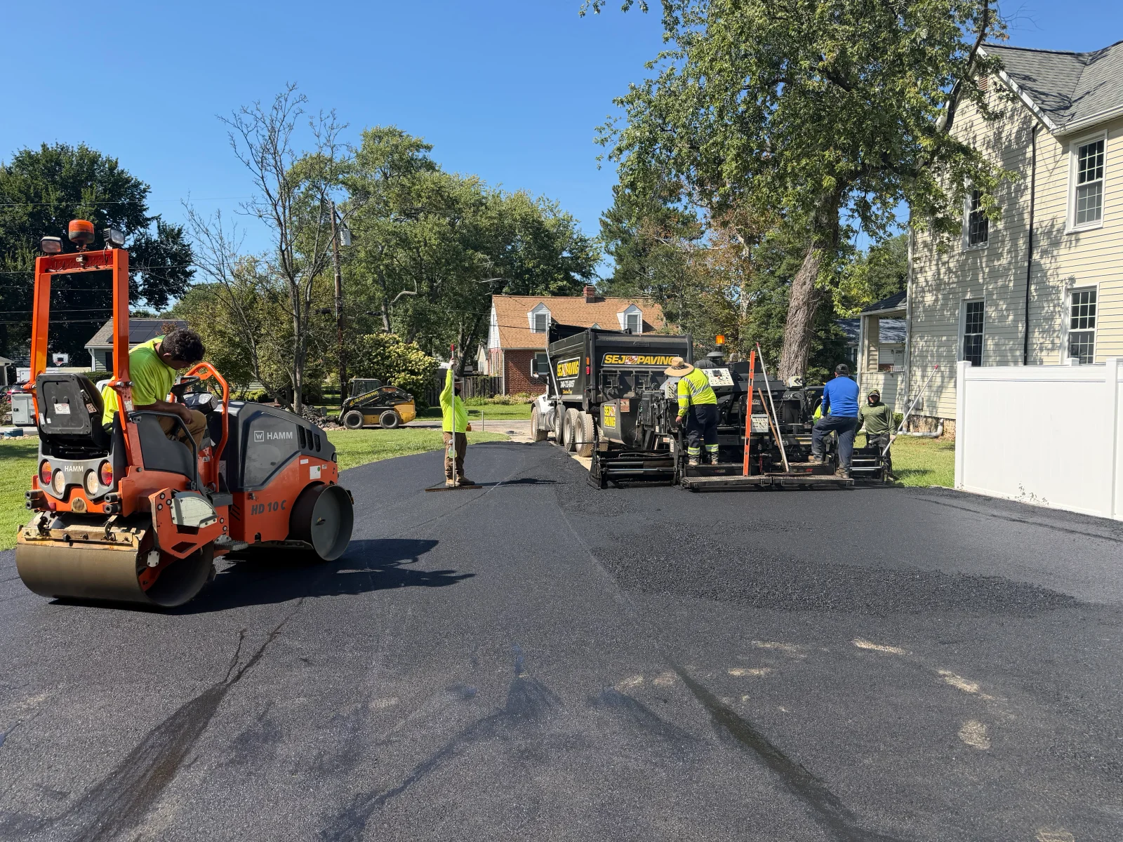 Crew laying hot-mix asphalt with paver, dump truck, and HAMM tandem roller on residential street
