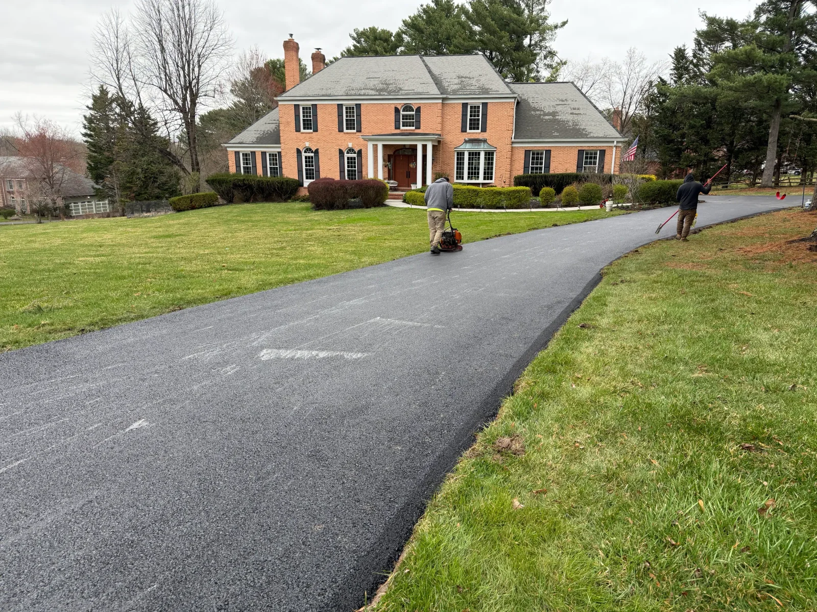 Freshly paved dark asphalt driveway curving past a lawn, with a crew member operating a plate compactor on the new surface