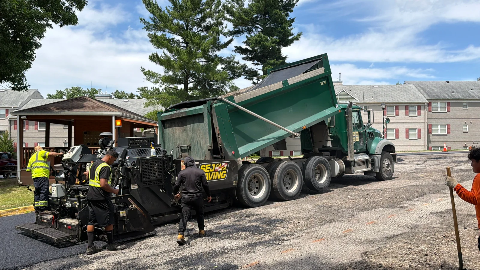 Paver laying fresh asphalt from green dump truck with crew in high-visibility vests on gravel base