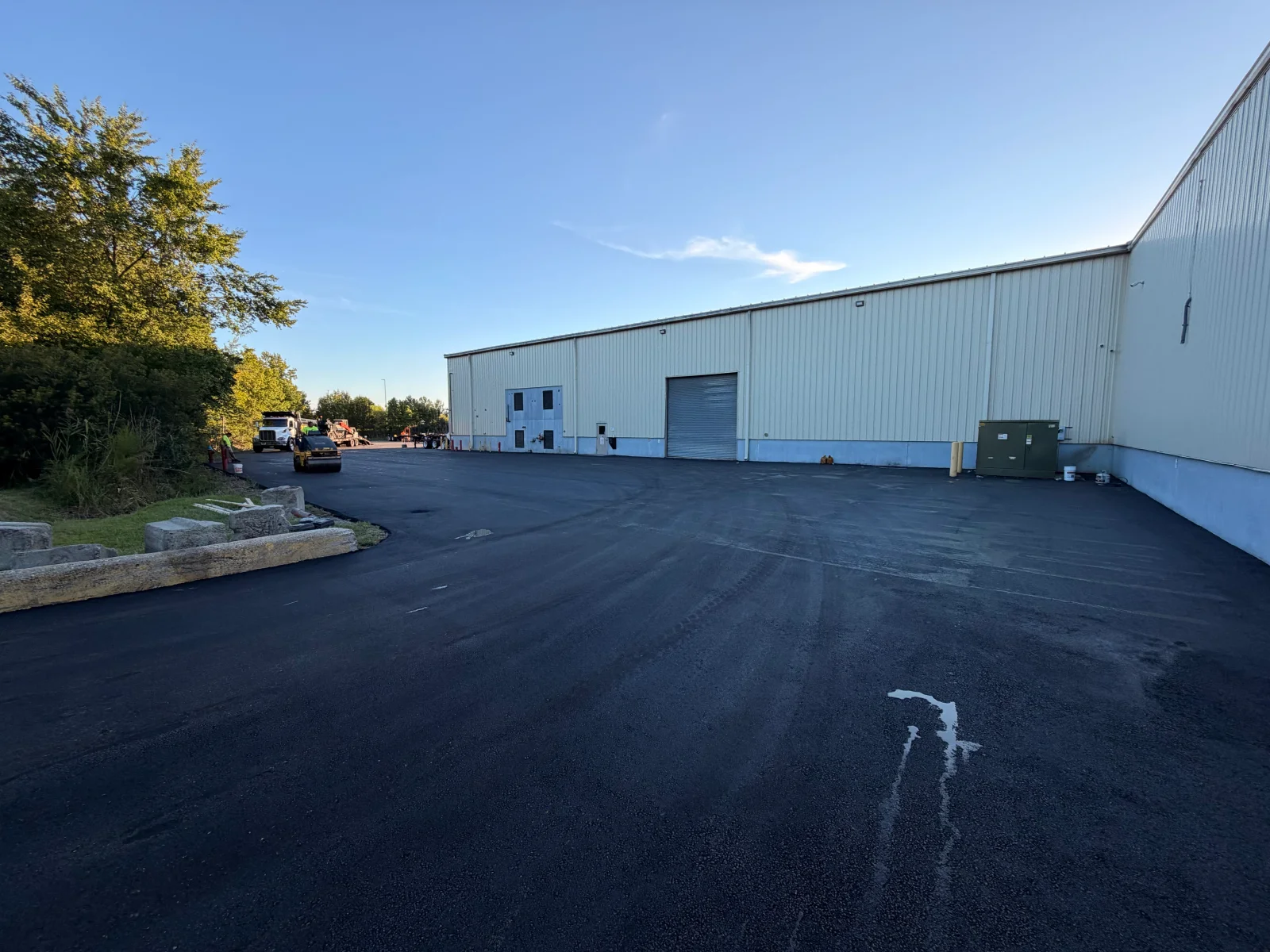 Newly paved black asphalt lot beside a metal warehouse with roll-up door, with roller and dump truck at work